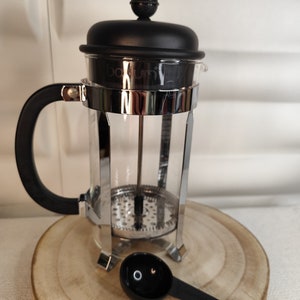 Peut inclure: Une cafetière à piston en verre noire et argentée avec une carafe en verre et un couvercle en plastique noir. La cafetière est posée sur une surface en bois. Une cuillère à mesurer en plastique noir se trouve devant la cafetière.
