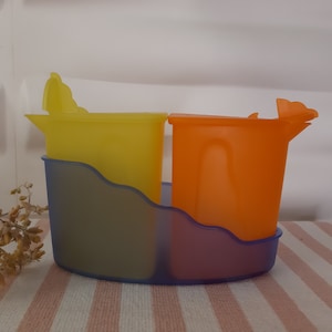 Op de afbeelding: Een bak met twee compartimenten met een blauwe basis en twee kleurrijke bekers. Eén beker is geel, de andere is oranje. Elke beker heeft een tuit en een deksel. De bak staat op een roze en wit gestreept oppervlak.