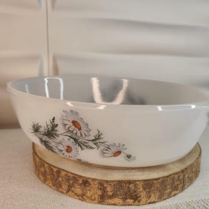 Puede incluir: Bol ovalado blanco con un diseño floral de margaritas. El bol tiene una superficie lisa y dos asas pequeñas. El diseño de la margarita presenta pétalos blancos y centros amarillos con hojas verdes. El bol está sobre una rodaja de madera.