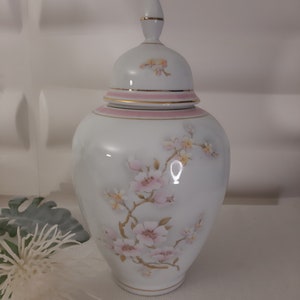 Può includere: Un vaso in porcellana bianca con un coperchio, decorato con fiori rosa e gialli. Il vaso ha un bordo dorato attorno al coperchio e alla base.