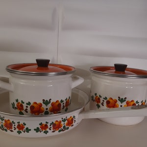 Pode incluir: Um conjunto de três panelas de esmalte branco com tampas laranja e desenhos florais. As panelas têm alças pretas e estão dispostas sobre uma superfície branca.