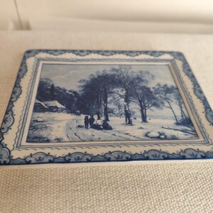 May include: A square ceramic tile with a winter landscape scene in shades of blue and white. The image depicts a snowy road, trees, and a building. The tile has a decorative border.