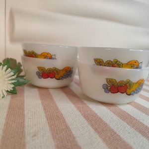 Puede incluir: Juego de cuatro cuencos blancos y redondos con un diseño de frutas colorido. Los cuencos presentan ilustraciones de fresas, uvas y naranjas. Los cuencos están apilados sobre una superficie a rayas.