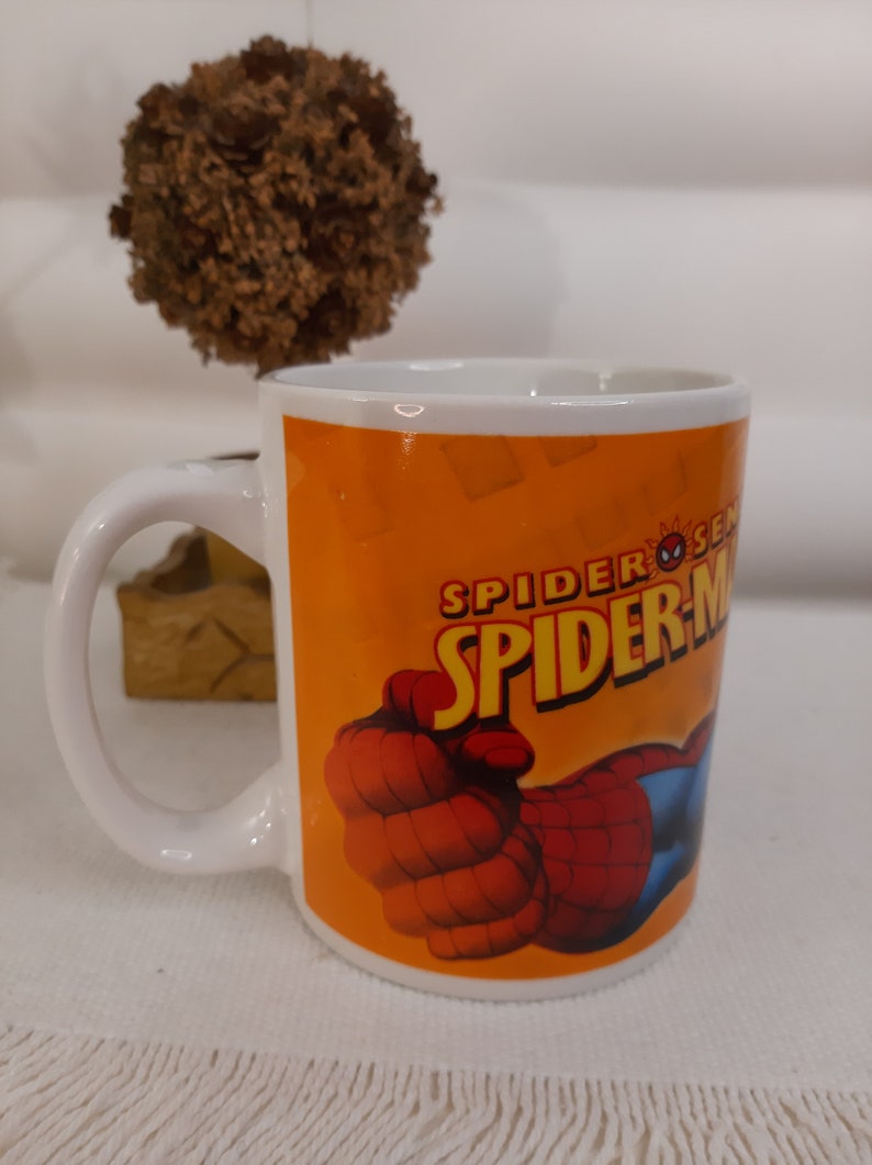 Op de afbeelding: Witte keramische mok met een feloranje achtergrond waarop een rode en blauwe Spiderman-illustratie is afgebeeld. De tekst "Spider-Sense" en "Spider-Man" is in zwart gedrukt.