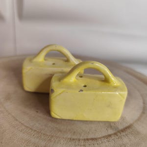 Op de afbeelding: Twee vintage gele zout- en peperstrooiers. Elke strooier heeft een rechthoekige vorm, een gebogen handvat en twee gaatjes. Ze staan op een houten oppervlak.