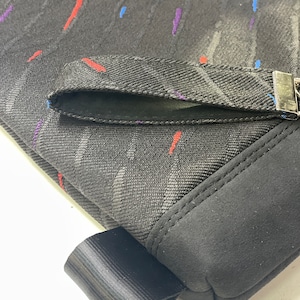 Könnte beinhalten: Schwarzer Stoff mit einer kleinen Tasche und einem schwarzen Riemen. Der Stoff hat ein dezentes Muster aus roten, blauen und violetten Linien.