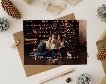 May include: A Christmas card featuring a family of three sitting in front of a decorated Christmas tree. The card has the text "Merry Christmas" in gold script and "Alicia, Nick & Olivia" in smaller text below.