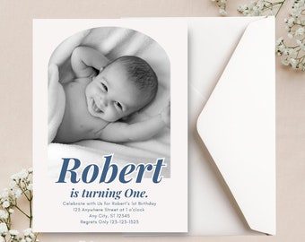 May include: A black and white photo of a baby boy on a white blanket. The photo is in a rounded frame with the text "Robert is turning One." below it. The text is in a blue font with a white outline.