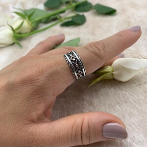 Pu&ograve; includere: Un anello d'argento con un delicato design floreale. L'anello &egrave; indossato su un dito e presenta un motivo ripetitivo di piccoli fiori.