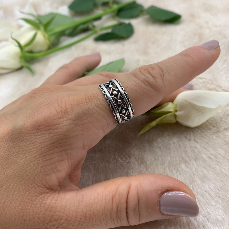 Pu&ograve; includere: Un anello d'argento con un delicato design floreale. L'anello &egrave; indossato su un dito e presenta un motivo ripetitivo di piccoli fiori.