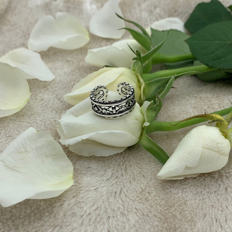 Pu&ograve; includere: Un anello per il dito del piede in argento con un design intricato, che presenta un delicato motivo a filigrana. L'anello &egrave; mostrato su uno sfondo bianco con petali di rosa bianca.