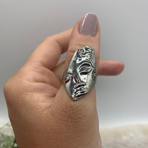 May include: A silver thumb ring with a detailed design of a face. The ring is worn on a person's thumb.