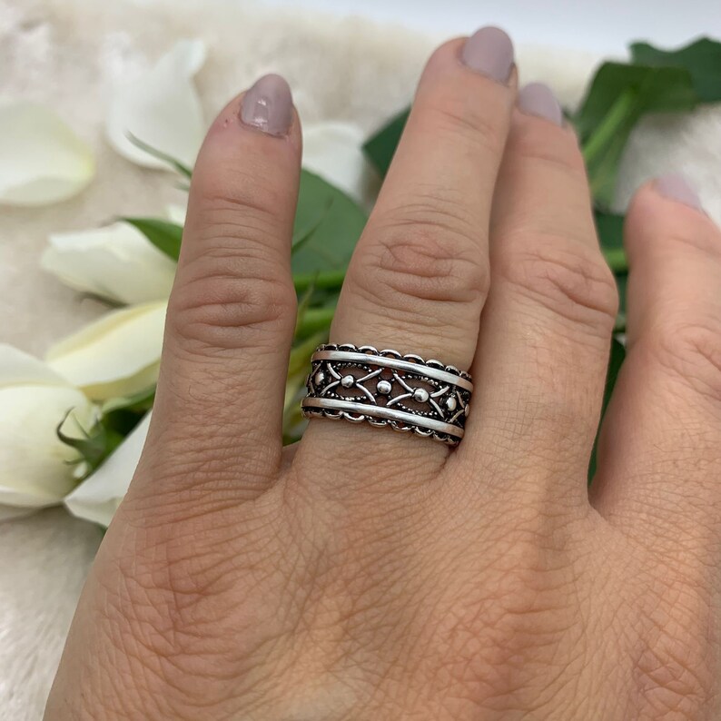 Pu&ograve; includere: Un anello d'argento con un design dettagliato e intricato. L'anello ha una fascia larga con un motivo ripetitivo di piccoli cerchi e diamanti.