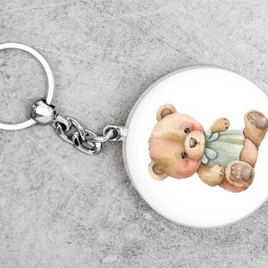 May include: A silver key chain with a round, white charm featuring a cartoon illustration of a brown teddy bear wearing a blue bow.