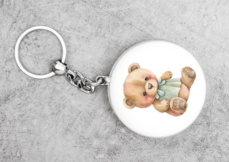 May include: A silver key chain with a round, white charm featuring a cartoon illustration of a brown teddy bear wearing a blue bow.