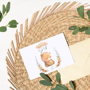 May include: A white card with a watercolor illustration of a brown bear sitting under a cloud with raindrops. The bear is surrounded by a wreath of leaves and the number 6 is below the bear.