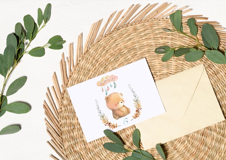 May include: A white card with a watercolor illustration of a brown bear sitting under a cloud with raindrops. The bear is surrounded by a wreath of leaves and the number 6 is below the bear.