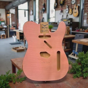 Puede incluir: Un cuerpo de guitarra rosa con secciones enrutadas para electrónica y un mástil. El cuerpo tiene una textura de veta de madera y una etiqueta que dice "White Stag" y el número "334530". El cuerpo de la guitarra está sobre una mesa de madera.