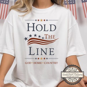 May include: A white short-sleeved t-shirt featuring the dark blue text HOLD THE LINE with a red wavy graphic and stars. Below, the text reads GOD HOME COUNTRY in red with small stars. The design includes a circular badge stating Comfort Colors.