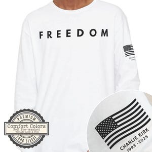 May include: White long-sleeve shirt with the word "FREEDOM" printed across the chest in black. Features a small American flag graphic on the right sleeve and a larger flag graphic with text. Includes a "Comfort Colors" logo.
