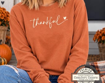 May include: A long-sleeve crewneck sweatshirt in a burnt orange color featuring the word thankful in white cursive script with a small heart icon at the end. The garment is made by Comfort Colors and has a vintage look and feel.