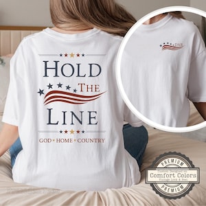May include: A white short-sleeve t-shirt featuring the text HOLD THE LINE in dark blue with a red wavy graphic and stars. The back displays the phrase GOD HOME COUNTRY with stars, while the front chest area shows a smaller matching graphic.