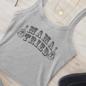 May include: A gray tank top with the text "MAMA TRIED" in a retro font, flanked by guitar illustrations. The top has a scoop neck and thin straps. The background includes a wooden surface and a gold necklace.