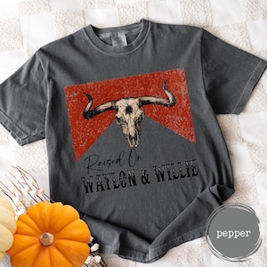May include: Dark grey t-shirt with a bull skull graphic and the text "Raised On Waylon & Willie" in black. The graphic is set against a red, distressed background. The shirt is a comfortable, casual style.