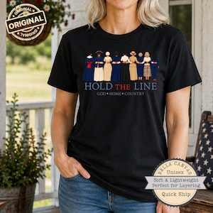 May include: A black t-shirt featuring a graphic of nine figures standing in a row with arms linked. Below the figures, the text reads HOLD THE LINE in blue and red, followed by GOD HOME COUNTRY in white. The shirt has short sleeves and a crew neck.