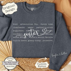 May include: A dark gray sweatshirt with the word "Sister" in cursive, surrounded by descriptive words like "kind" and "supportive." The sleeve features the name "baylee billie." Includes a "Custom Names" option.