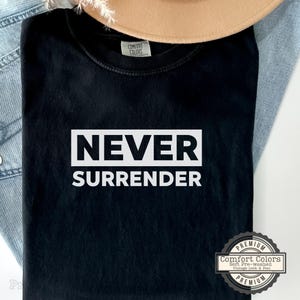 May include: Black t-shirt with the words "NEVER SURRENDER" in a white rectangular box. The shirt has a vintage look and feel, with a Comfort Colors label. A tan hat and blue jeans are in the background.