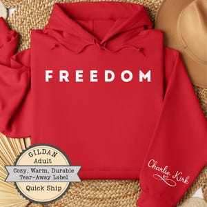 May include: A red hoodie with the word "FREEDOM" printed in white capital letters across the chest. The sleeve features the name "Charlie Kirk" in white script. The sweatshirt has a "GILDAN Adult" label.