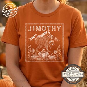 May include: A burnt orange short-sleeve t-shirt featuring a white graphic print of a raccoon walking in front of mountains and three pumpkins. The text JIMOTHY is printed in a white serif font above the raccoon within a square border.