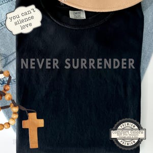 May include: Black t-shirt with the phrase "NEVER SURRENDER" in gray block letters. The shirt also features the text "you can't silence love" on a white sticker. A wooden cross and a string of wooden beads are visible.