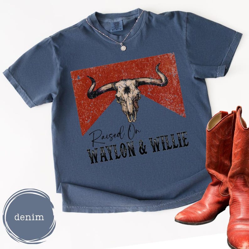 May include: A denim blue t-shirt featuring a bull skull graphic on a red backdrop, with the text "Raised On Waylon & Willie" beneath. A pair of red cowboy boots are positioned to the right of the shirt.