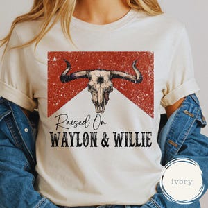 May include: Cream-coloured t-shirt with a distressed graphic of a bull skull over a red and white background. The text "Raised On WAYLON & WILLIE" is printed below the graphic in a bold, black font. The shirt has short sleeves.