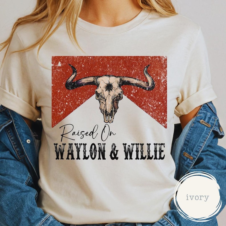 May include: Cream-coloured t-shirt with a distressed graphic of a bull skull over a red and white background. The text "Raised On WAYLON & WILLIE" is printed below the graphic in a bold, black font. The shirt has short sleeves.