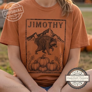 May include: A burnt orange short-sleeve t-shirt featuring a black graphic print of a raccoon walking in front of a mountain range and three pumpkins. The text JIMOTHY is printed in a bold, decorative font above the mountain illustration.