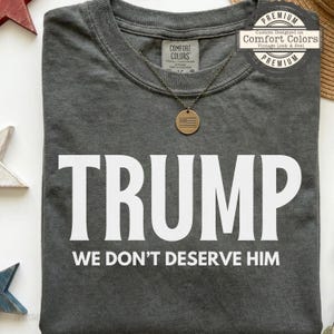 May include: A charcoal grey t-shirt featuring the white text TRUMP WE DON'T DESERVE HIM printed across the chest. A circular pendant necklace with an American flag design rests on the neckline. The shirt has a Comfort Colors brand label.
