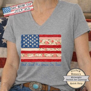Op de afbeelding: Een grijs V-hals T-shirt met een patchwork Amerikaans vlagontwerp. De vlag bevat bloemen- en ruitpatronen in rood, wit en blauw. De tekst "God Shed His Grace On Thee" is eronder gedrukt. Het shirt heeft de tekst "Get NOW for JULY 4th".