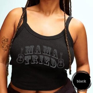 May include: Black tank top with the text "MAMA TRIED" in a distressed font, accompanied by guitar illustrations. The top has a scoop neckline and wide straps. The word "black" is in a circle on the bottom right.
