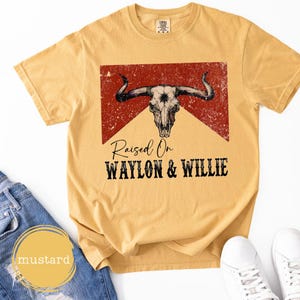 May include: Mustard yellow t-shirt with a distressed bull skull graphic and the text "Raised On Waylon & Willie". The shirt has a crew neck and short sleeves. The shirt is made of a soft, comfortable material. The shirt is a casual wear item.