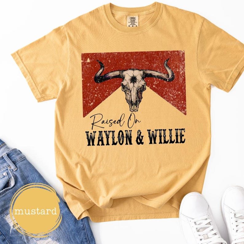 May include: Mustard yellow t-shirt with a distressed bull skull graphic and the text "Raised On Waylon & Willie". The shirt has a crew neck and short sleeves. The shirt is made of a soft, comfortable material. The shirt is a casual wear item.
