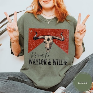 May include: A moss green t-shirt featuring a distressed bull skull graphic on a red backdrop. The text "Raised On Waylon & Willie" is printed below. The shirt has a relaxed fit and is a casual style.