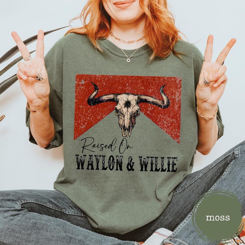 May include: A moss green t-shirt featuring a distressed bull skull graphic on a red backdrop. The text "Raised On Waylon & Willie" is printed below. The shirt has a relaxed fit and is a casual style.
