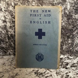 May include: A vintage book titled "THE NEW FIRST AID IN ENGLISH" with a blue cover and a dark blue cross symbol. The author's name, Angus Maciver, is printed below the cross. A small emblem is at the bottom.