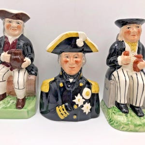 May include: Three ceramic figurines of historical figures. The figures wear period clothing, including tricorn hats and military uniforms. Each figure holds a small jug or bottle. The figurines are set on a green base.