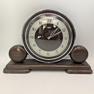 May include: A dark brown wooden mantel clock with a circular face, a silver-toned metal bezel, and a glass cover. The clock features a white dial with black Arabic numerals from 1 to 12, two silver-toned hands, and a base with two spheres.