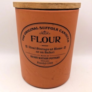 May include: A terracotta-colored flour canister with a wooden lid. The canister features a black circular label with the text "The Original Suffolk Canister FLOUR" and "Ideal Storage at Home or on Safari". The label also includes "HENRY WATSON POTTERY ENGLAND Founded 1800".
