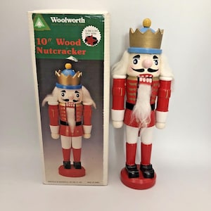 May include: A 25.4 cm wooden nutcracker figure wearing a gold crown, blue hat, and red uniform with white accents, featuring a white faux fur beard and hair. It stands beside its original rectangular cardboard box labelled Woolworth 10" Wood Nutcracker.